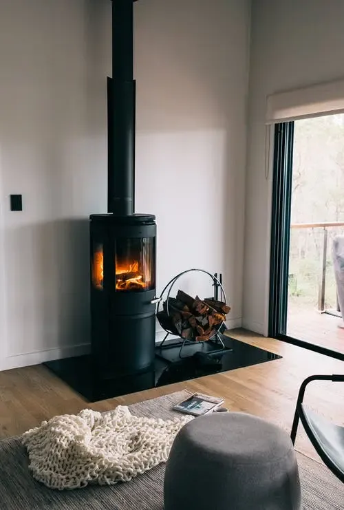 Angolo relax con stufa a legna e vista sul balcone Interno accogliente con stufa a legna nera, legna accatastata, tappeto intrecciato e vista su un balcone attraverso una porta scorrevole in vetro